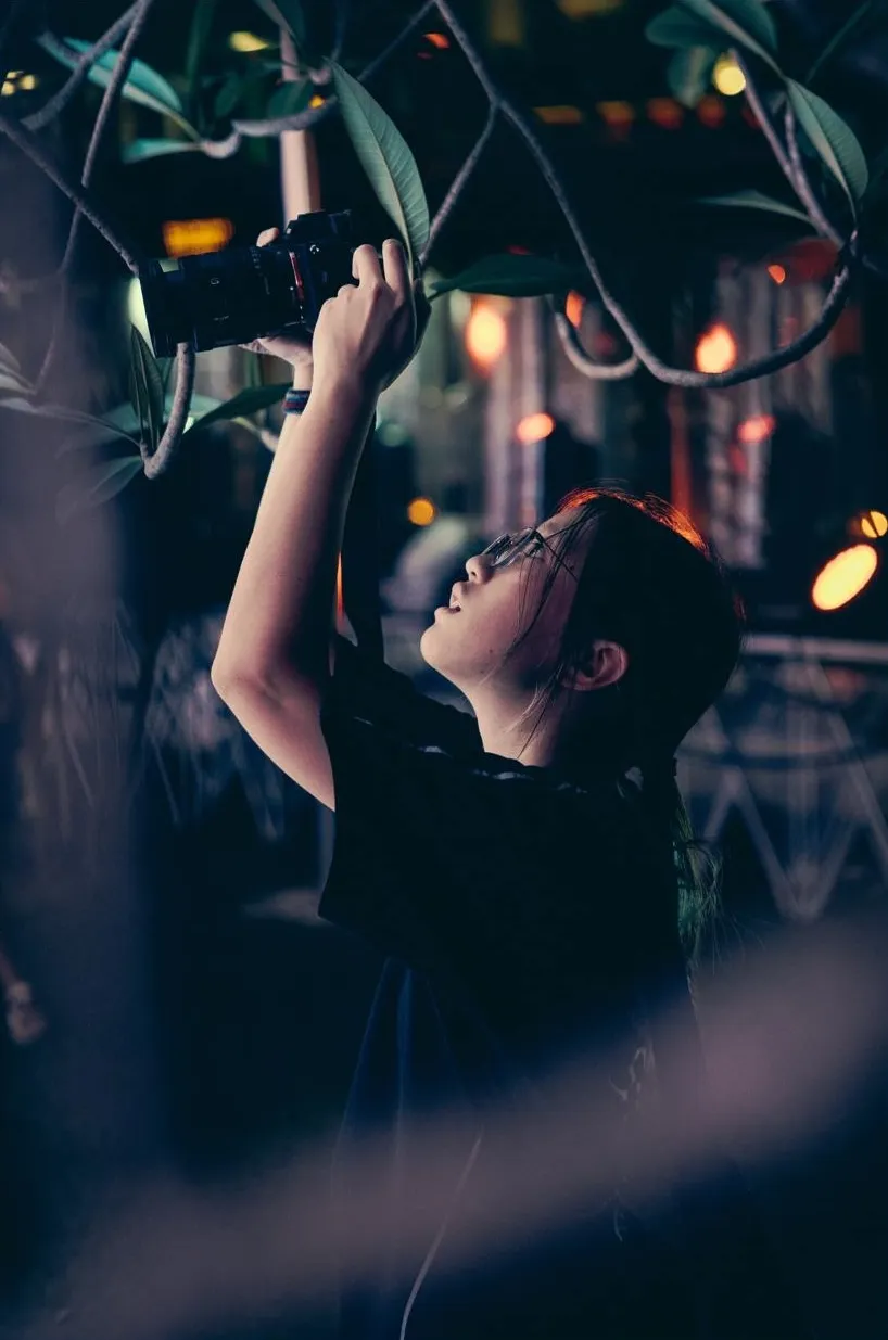 Tessa Leong holding her camera up high among leaves with beautiful night city lights in the background.